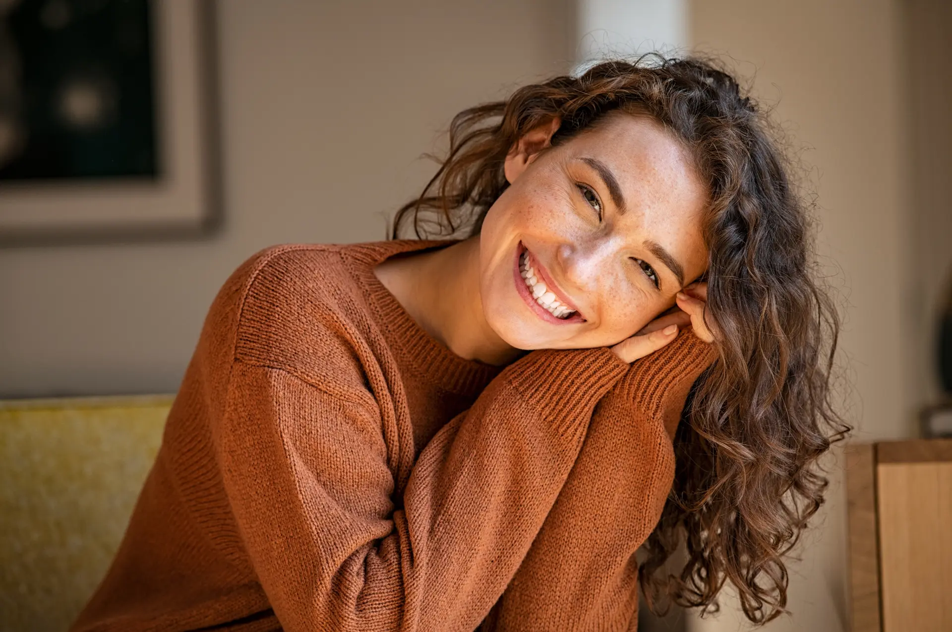Smiling woman in a warm rust knit sweater resting her head on her arms in a relaxed home setting